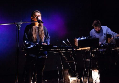 Image of the musical duo Hyperdawn, a man and a woman stood next to each other. The woman is turning dials on a synthesiser and singing into a microphone. The man is also turning dials on a synthesiser.