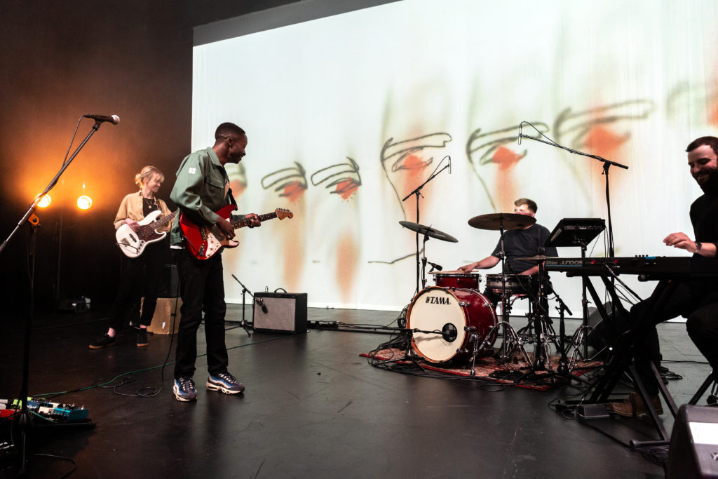 Image of the band Nii performing onstage at Art School Live. Consists of a woman playing bass, a man playing guitar and singing, a man playing the drums, and a man sat playing the keyboard.