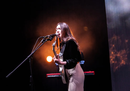 Image of Natalie McCool stood playing a white guitar and singing into a microphone.